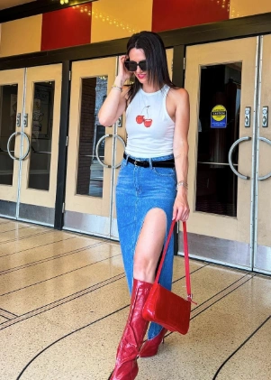 A woman in a white sleeveless top with cherry prints paired with a denim skirt with a thigh-high slit and red knee-high boots, accessorized with a red handbag, giving off an edgy yet fashionable brunch vibe.