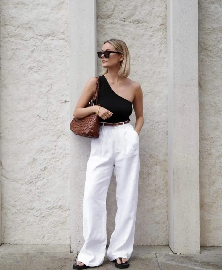 A woman in a one-shoulder black top and white high-waisted trousers with black sandals and a woven brown bag — European summer style.