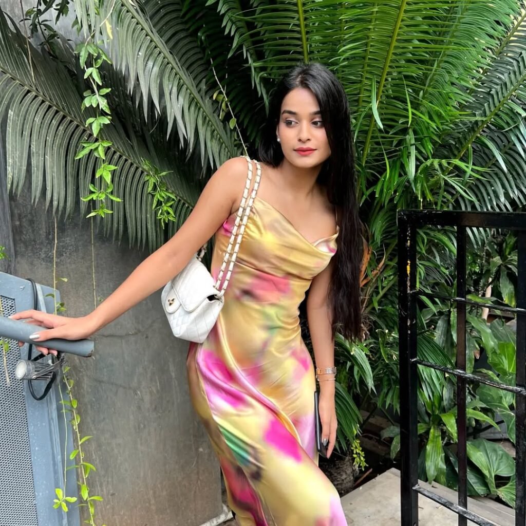 A woman in a satin multicolored dress, featuring a blend of yellow, pink, and green hues, paired with a white handbag and standing near greenery, exuding a relaxed yet chic vibe.