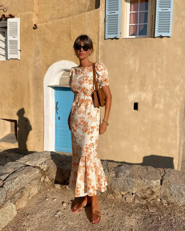 A woman in a fitted floral midi dress with puff sleeves and tan sandals, standing by a sunlit stone wall — a romantic and vintage-inspired European summer outfit.