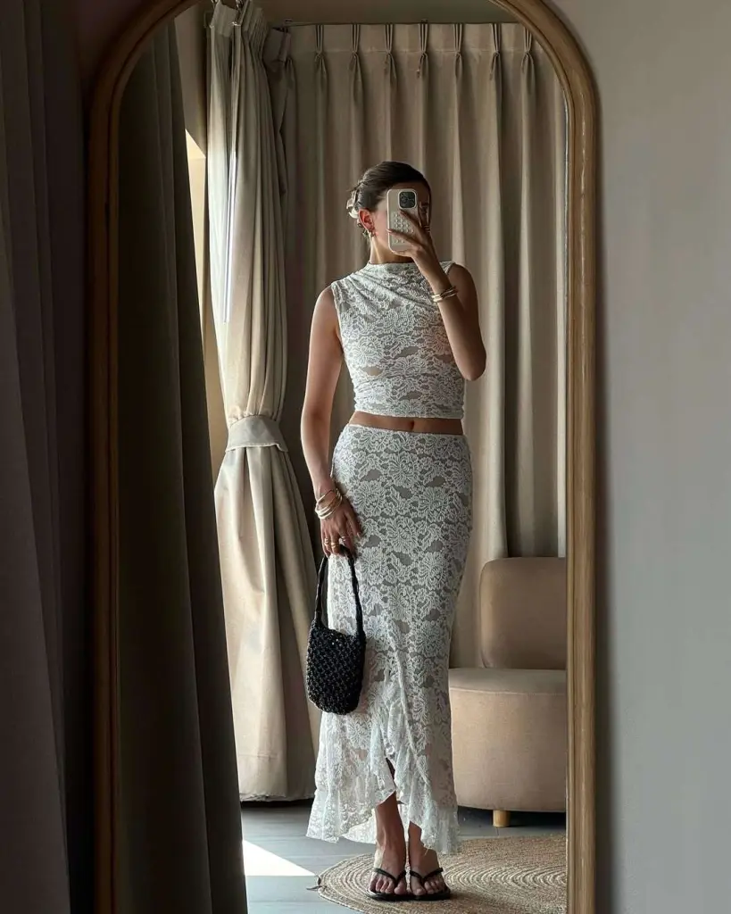 Beach outfit in a white lace crop top and matching asymmetrical lace skirt, accessorized with black woven handbag and flip-flops, captured in a mirror selfie.