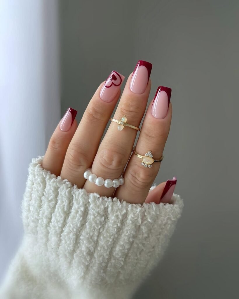 Red Nail Ideas showcasing a burgundy French tip manicure with a heart accent nail, complemented by pearl and gemstone rings.