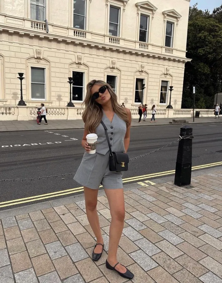 A woman sipping coffee in a tailored grey vest and shorts set with black ballet flats and a crossbody bag — a stylish business-casual European summer outfit.