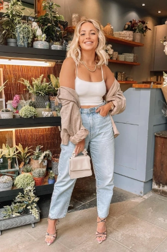 A woman in a white tank top and light blue jeans, paired with a beige cardigan and heels, holding a small handbag, showcasing a casual and stylish brunch outfit ideal for a summer day out.