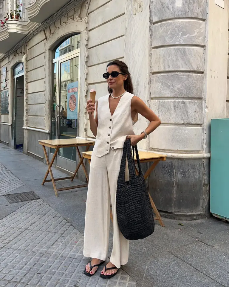 A woman eating ice cream in a sleeveless cream linen set with wide-leg trousers and a large black woven bag — a breezy and elegant European summer outfit.