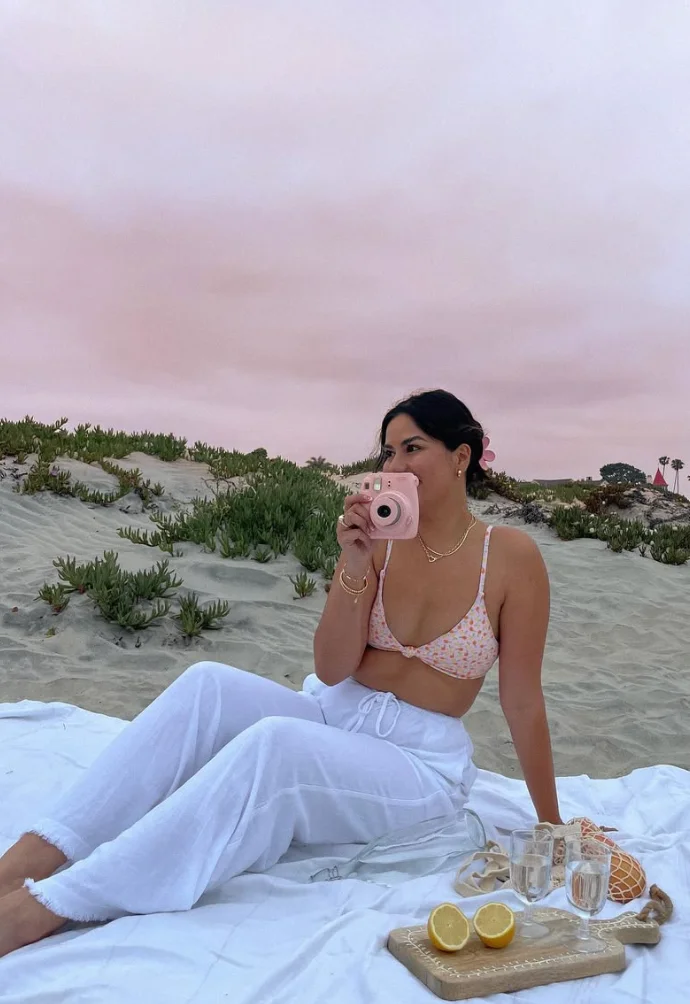 Beach outfit with a pink floral bikini top and loose white linen pants, accessorized with minimal gold jewelry, lounging on a beach picnic setup at sunset.