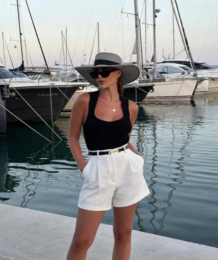 A woman in a black tank top tucked into high-waisted white shorts, accessorized with a striped wide-brim hat and dark sunglasses by a marina — a chic nautical European summer outfit.