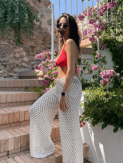Woman in white open-knit crochet pants and a red bikini top, posing on outdoor stairs with flowers in the background – Crochet Outfits.

