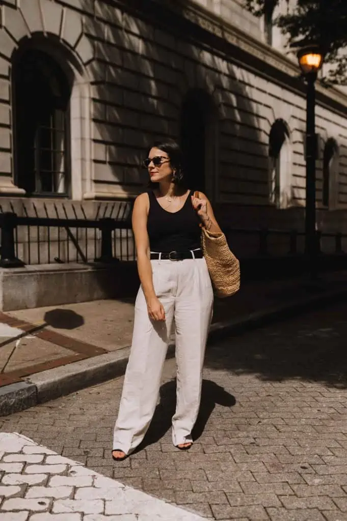 Woman in white wide-leg linen pants and a black tank top, with a woven bag and sandals. Clean, casual linen pant outfit.