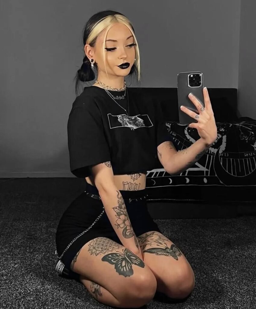 Grunge Outfits: Person with black and blonde hair styled in pigtails, wearing a black crop top and black shorts with tattoos on arms and legs, taking a mirror selfie.