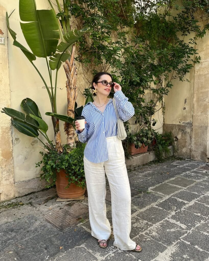 Casual summer look with white linen pants and a blue striped button-up shirt, accessorized with black sunglasses and a handbag.
