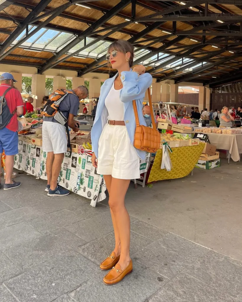 Woman in a light blue blazer over a white tank top and white shorts, paired with tan loafers and a woven handbag, shopping at a local market — european summer outfits.
