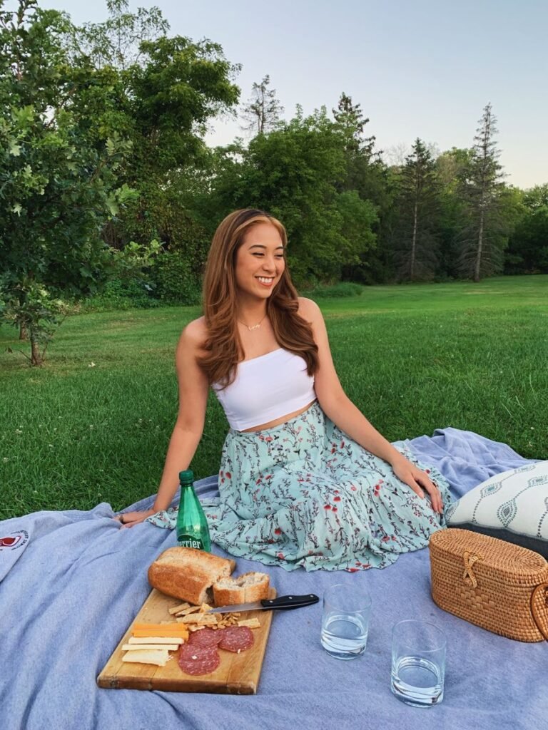 Picnic Date Outfits: Smiling woman in a white top and floral skirt sitting on a blanket with a charcuterie board and Perrier.