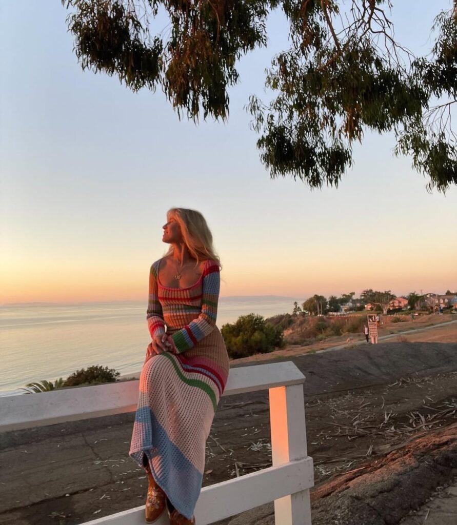 Woman sitting on a fence at sunset, wearing a long-sleeve multicolor striped crochet dress – Crochet Outfits.