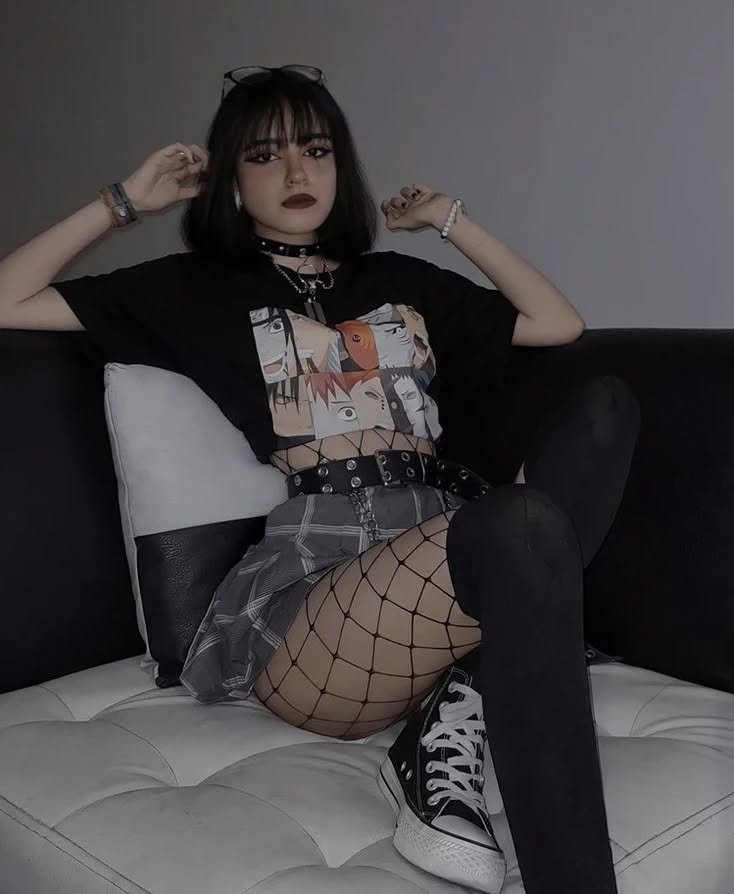 Grunge Outfits: Person with short black hair wearing a black choker, black graphic t-shirt, plaid skirt, fishnet stockings, and black sneakers sitting on a couch.