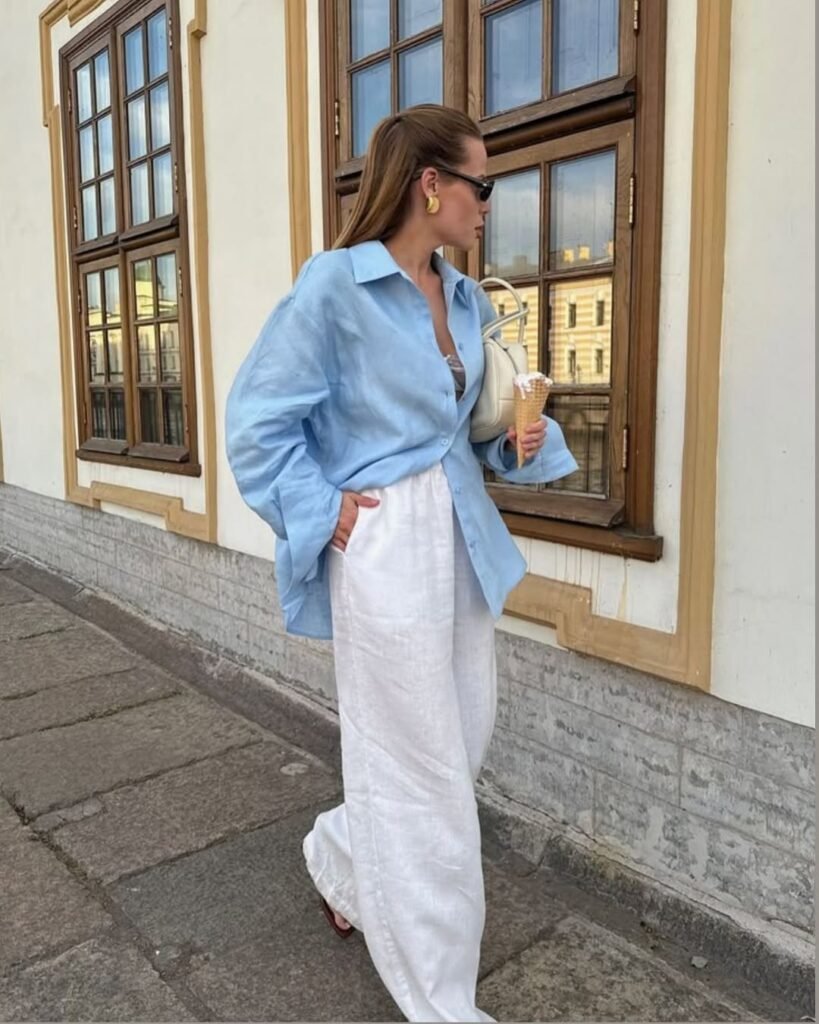 Stylish woman wearing white wide-leg linen pants and a light blue shirt, holding an ice cream cone while walking on a sidewalk.