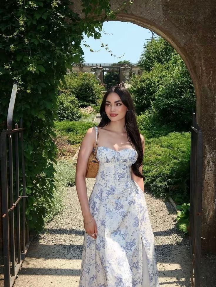 A woman in a satin floral dress with a sweetheart neckline, walking through a lush garden, carrying a small handbag.