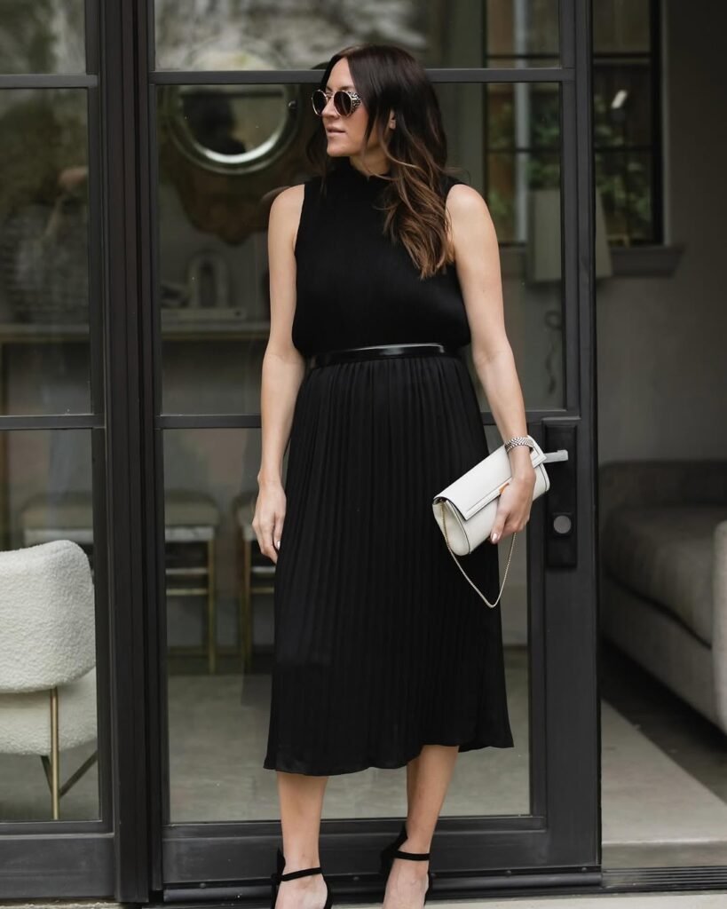 Stylish work attire in an all-black pleated midi dress with a high neckline, accessorized with a white clutch and black heels for a sleek and elegant office outfit.