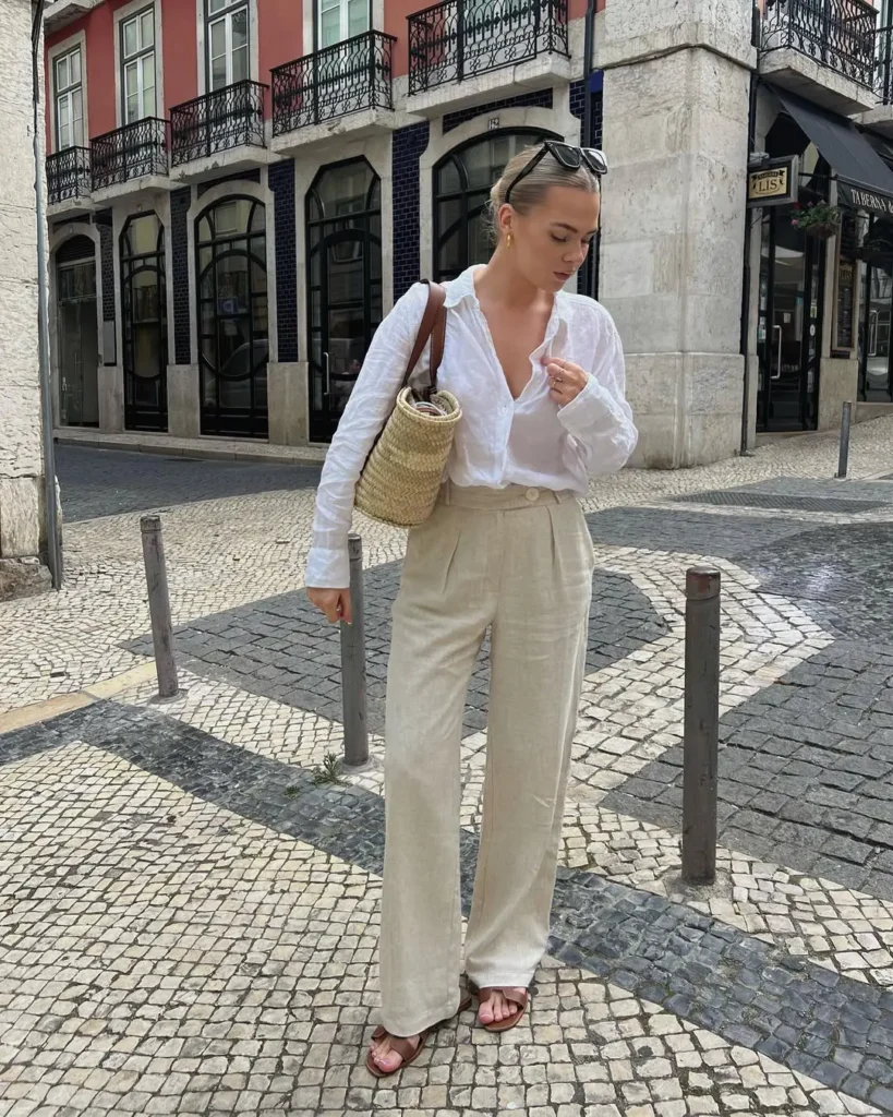 A woman in a white linen shirt and beige wide-leg trousers with flat sandals, carrying a straw bucket bag — European summer style.