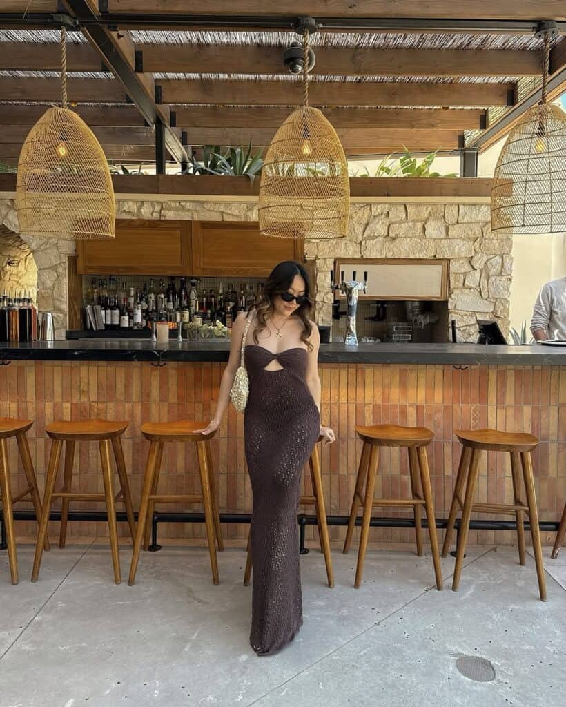 Beach outfit with a brown crochet strapless maxi dress featuring a center cut-out, paired with a pearl handbag and dark sunglasses at a beach bar.