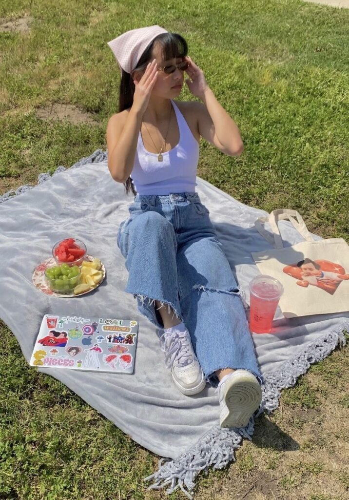 Picnic Date Outfits: Woman in a white tank top, jeans, and headscarf enjoying a casual picnic with fruit and a pink drink.