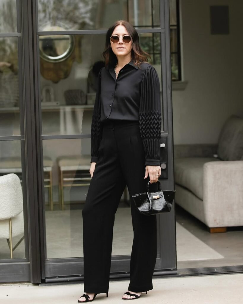 Stylish work attire featuring a black blouse with pleated sleeves and wide-leg trousers, paired with a mini black handbag and strappy sandals for a sophisticated look.