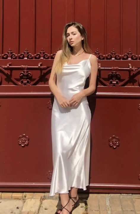 A woman in a satin white dress with spaghetti straps, posing near a red gate, exuding a calm and confident aura.