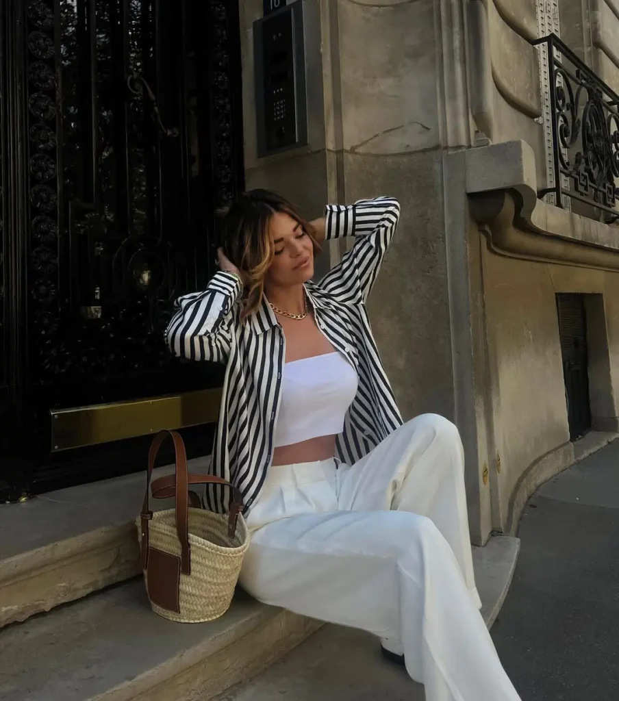 A woman in white wide-leg pants, a white crop top, and a black and white striped shirt, sitting on steps with a straw handbag — European summer style.