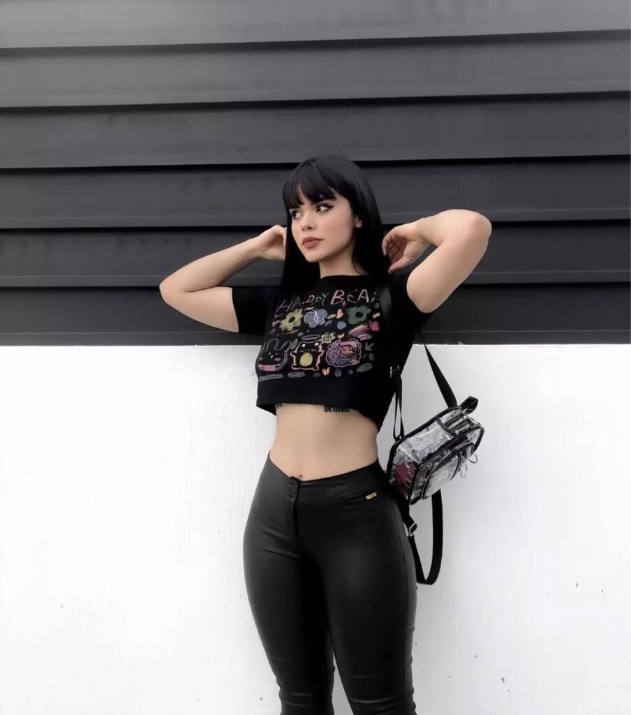 Grunge Outfits: Person with long dark hair wearing a black crop top with reflective lines and black high-waisted pants posing outdoors in sunlight.