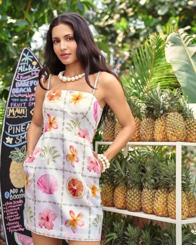 Woman wearing a sleeveless white dress with tropical fruit and flower prints, accessorized with pearl jewelry, ideal for fresh outfits verano.