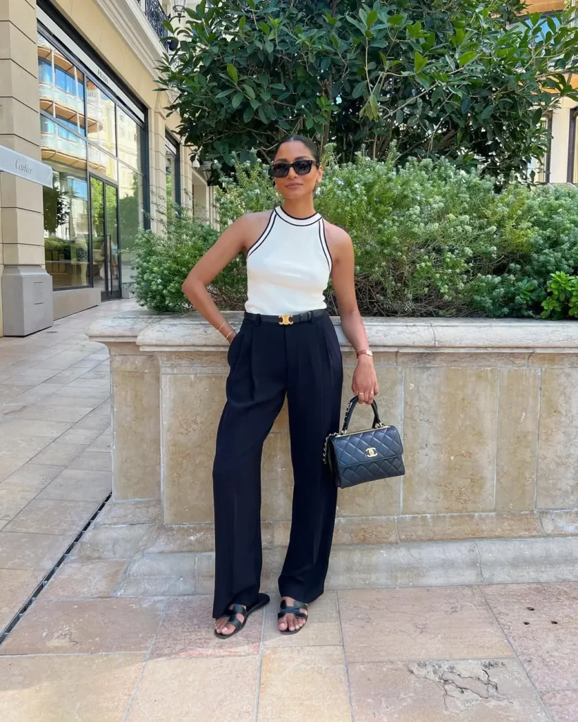 A woman in black tailored trousers and a sleeveless white top with black trim, holding a black quilted handbag — European summer style.