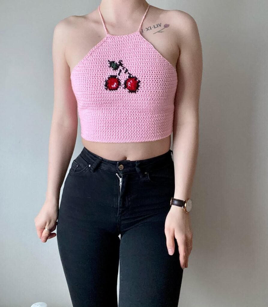 Woman dressed in a pink halter crochet crop top featuring a cherry design, paired with high-waisted black pants – Crochet Outfits.