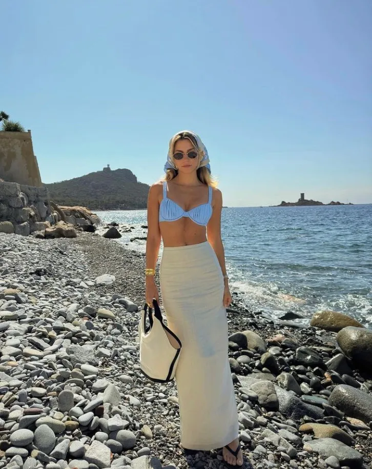 Beach outfit including a pastel blue bikini top with a long cream skirt, headscarf, and round sunglasses, captured on a rocky shoreline.