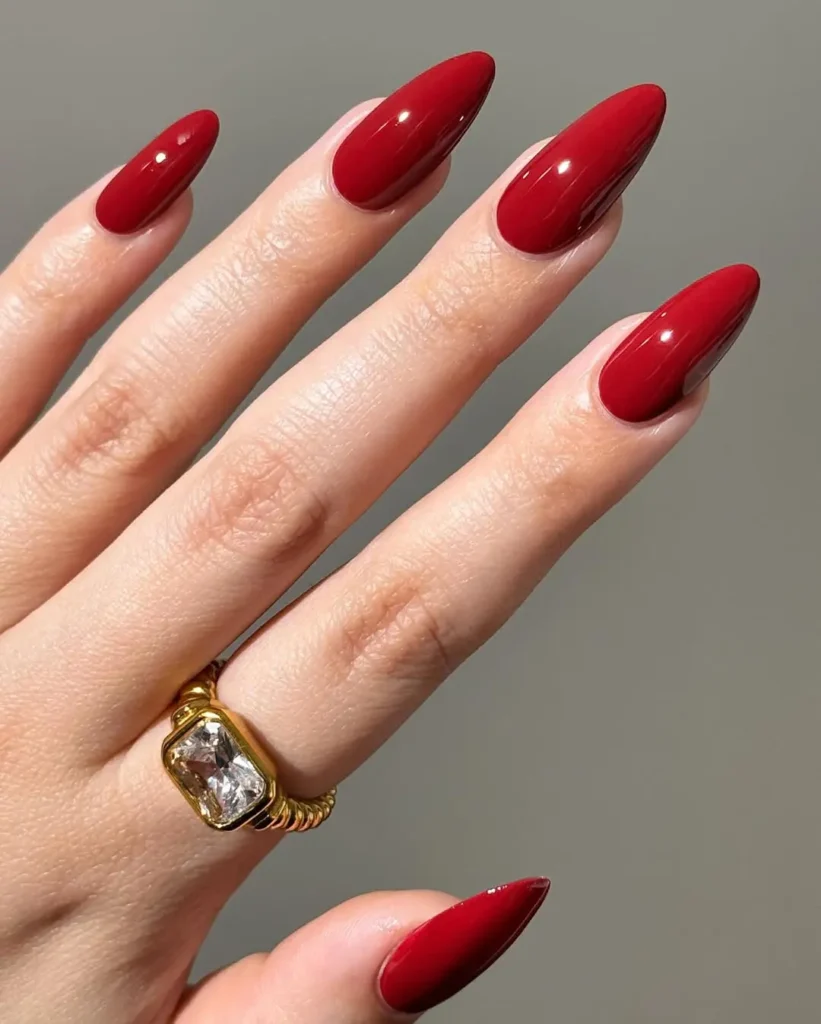 Glossy deep red almond-shaped nails paired with a large statement gold ring, bold Red Nail Ideas.