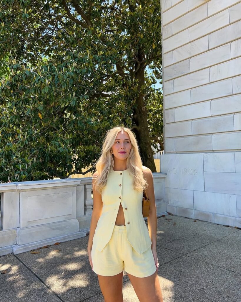 A woman in a yellow brunch outfit for summer, consisting of a sleeveless vest and shorts, standing outdoors with a natural tree backdrop.


