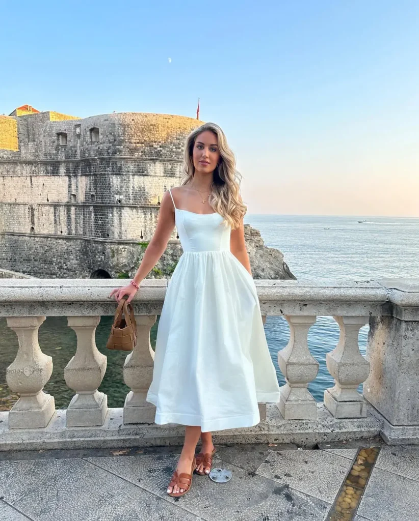 A woman in a white sundress with spaghetti straps and brown sandals, posing by the sea with a brown handbag — European summer style.