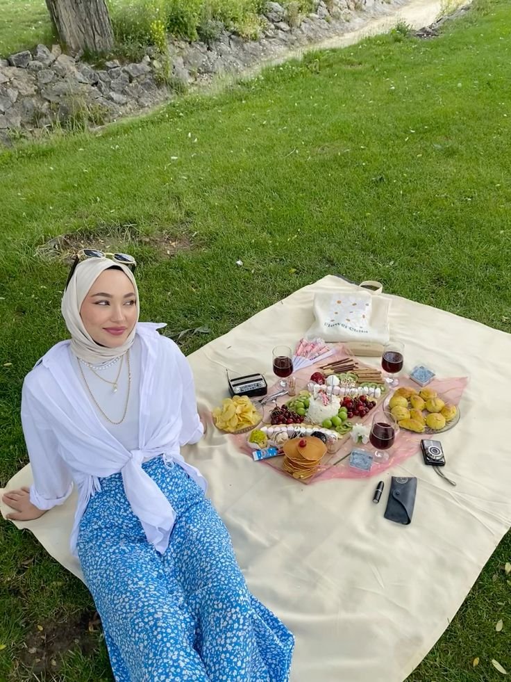 Picnic Date Outfits: Woman in a blue floral skirt, white top, and beige hijab relaxing at a scenic picnic with various snacks.