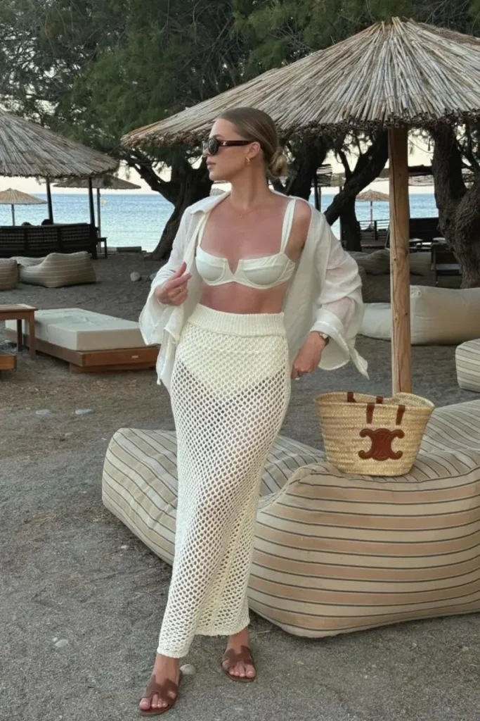 Beach outfit with a white bikini top and a cream crochet maxi skirt, paired with an oversized white shirt and straw handbag, perfect for a seaside resort.