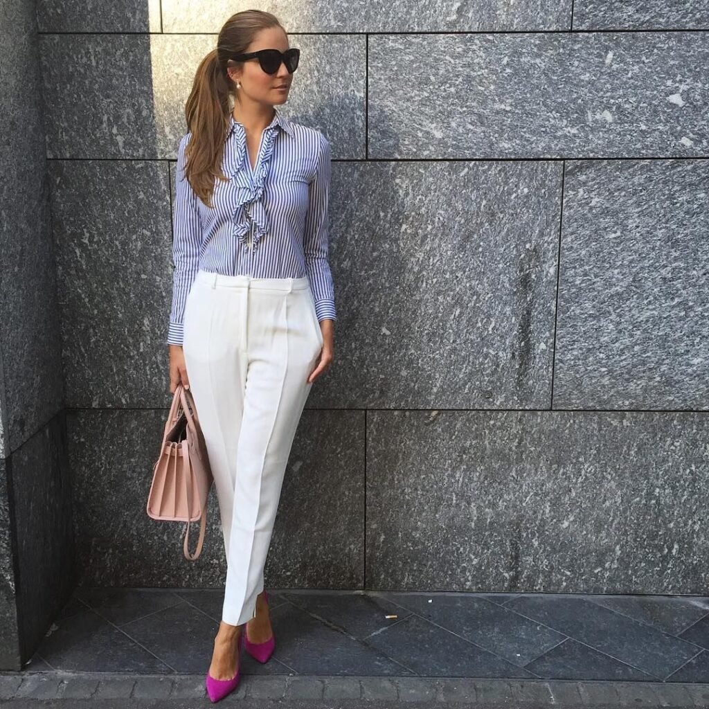 Stylish work attire with a blue striped ruffle blouse tucked into high-waisted white trousers, finished with bright pink heels and a blush handbag for a chic pop of color.