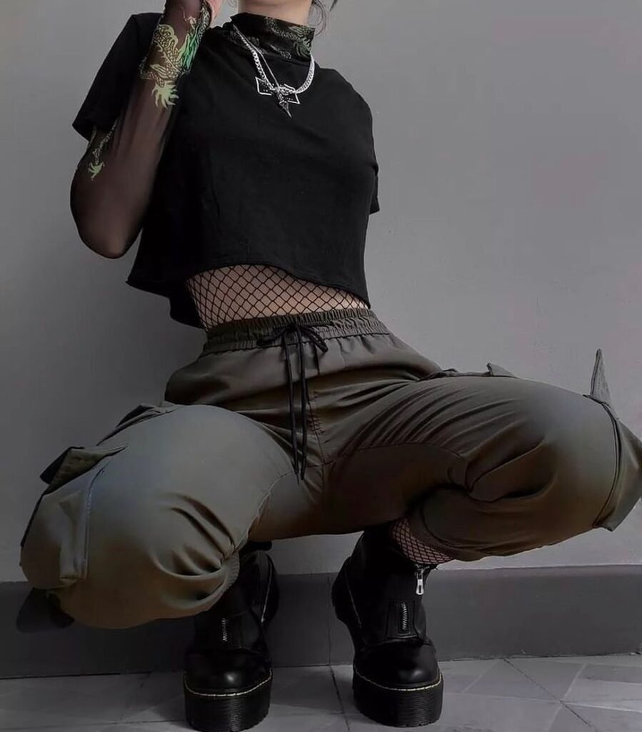 Grunge Outfits: Person wearing a black long sleeve crop top with buckles and ripped jeans over fishnet tights, sitting on stairs with black sneakers.