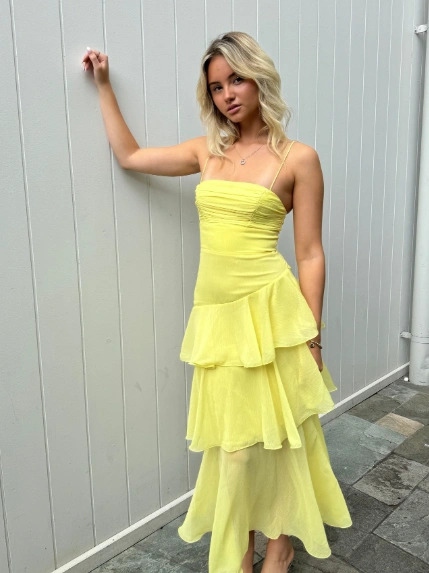 A woman in a strapless yellow tiered midi dress posing against a white wall; soft and feminine summer outfit idea.