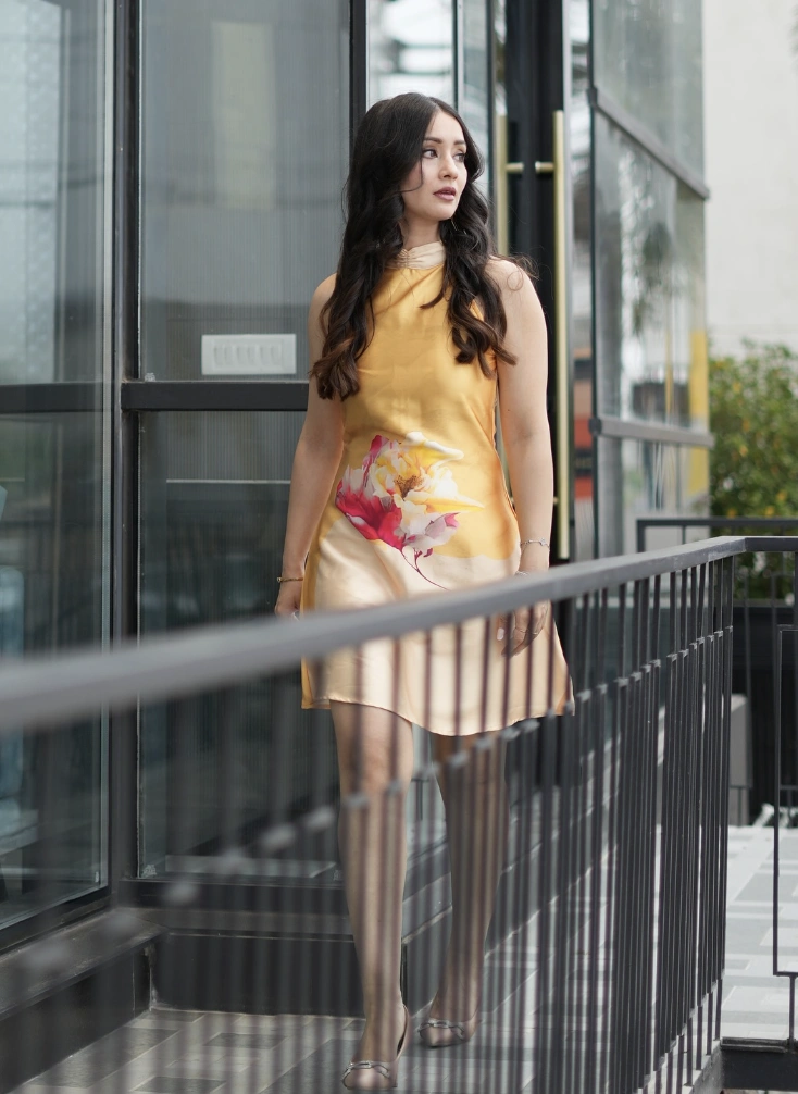 A woman in a satin yellow dress with floral prints, walking on a balcony in a modern urban setting, exuding elegance.