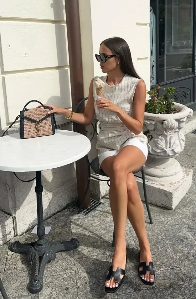 A woman seated at an outdoor café in a sleeveless beige pinstripe romper with black sandals and a structured straw handbag — European summer style.