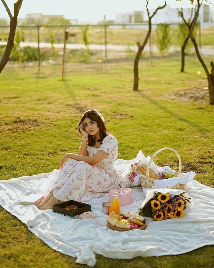 Picnic Date Outfits: Woman in a floral white dress enjoying a sunny picnic with cake, juice, and sunflowers on a blanket.