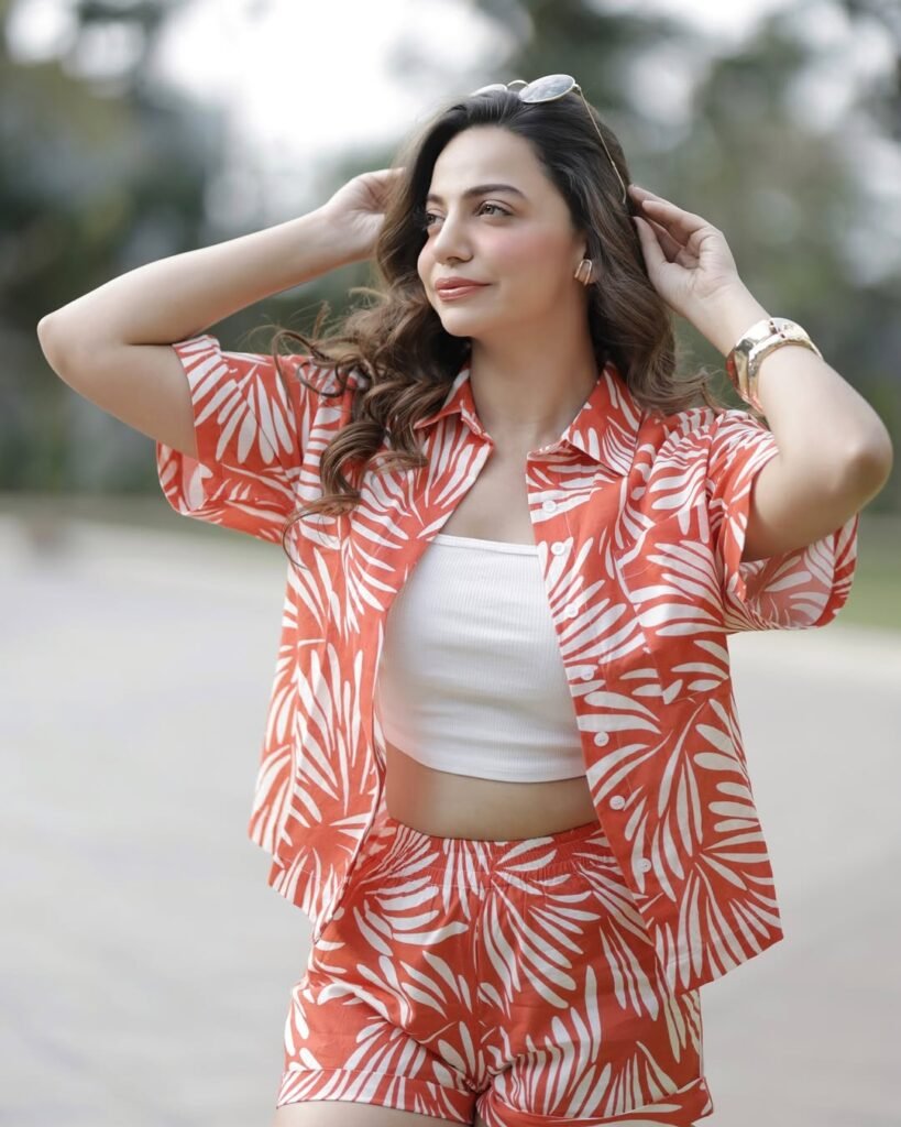 A woman in an orange and white printed summer brunch outfit, featuring a loose-fitting shirt and matching shorts, paired with sunglasses and posing outdoors.