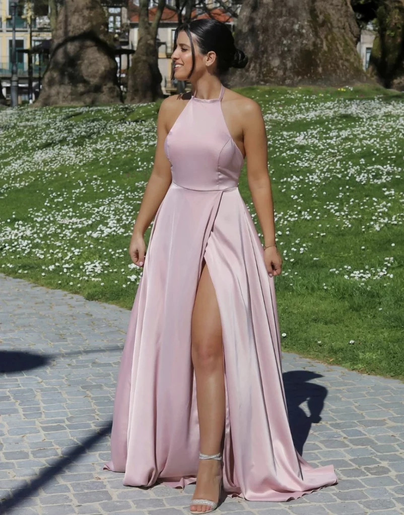 A woman in a satin pink dress with a thigh-high slit, walking outdoors in a natural setting, smiling with a fresh and joyful expression.