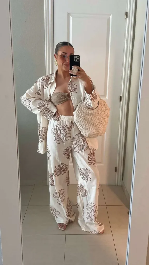 Beach outfit with a matching tropical print oversized shirt and trousers, worn over a bandeau bikini top, accessorized with a woven tote bag and large hoop earrings.

