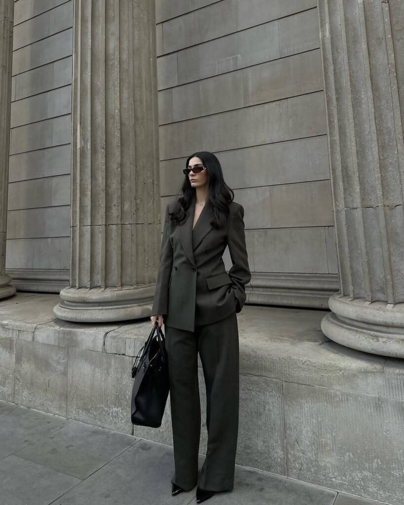 Stylish work attire in a dark olive oversized pantsuit with a double-breasted blazer and pointed heels, accessorized with a black leather handbag for a bold business presence.