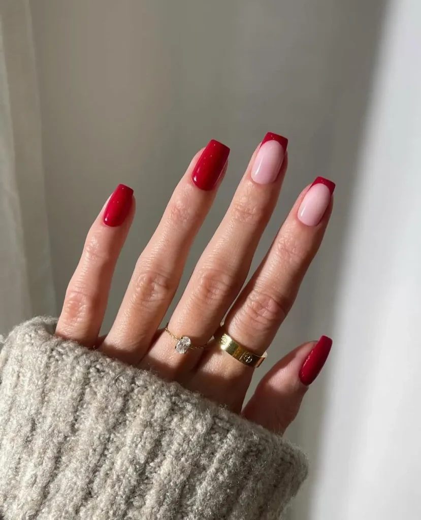A close-up of a hand with almond-shaped nails, painted with a glossy deep red polish and nude accent nails outlined with red, representing stylish Red Nail Ideas.