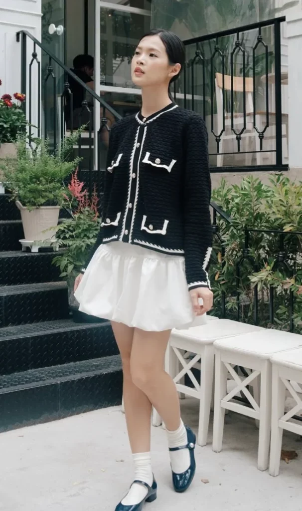 Woman wearing a black textured cardigan paired with a white pleated mini skirt, complemented by white socks and black Mary Jane shoes, standing outdoors by a garden area.







